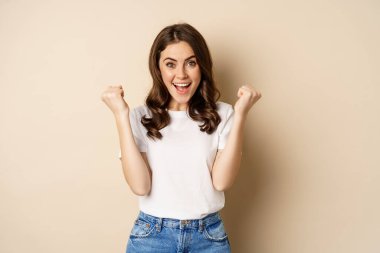Enthusiastic woman winning and fist pump, celebrating victory, smiling excited, standing over beige background
