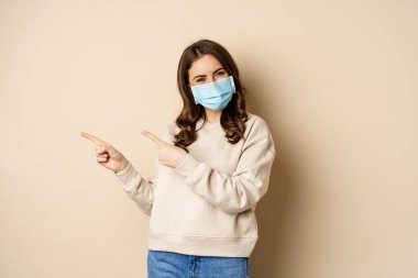 Health and covid-19 concept. Confused young woman in medical face mask, pointing left and staring with frustrated face, beige background