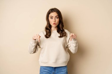 Enthusiastic attractive girl pointing fingers down, showing advertisement, link or promo below, standing over beige background