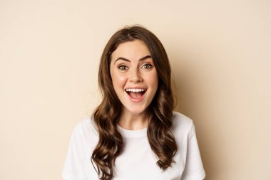 Close up portrait of surprised and excited brunette girl looking amazed, reacting impressed and excited, standing against beige background