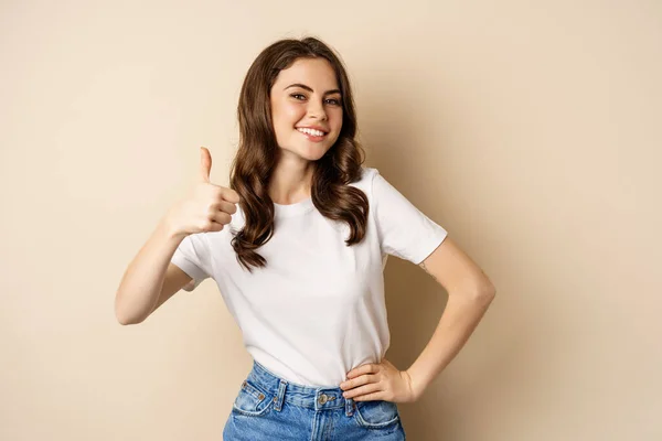 Portrait of modern young woman showing thumbs up, like and approve, smiling pleased, recommending company or website, standing over beige background — Fotografia de Stock