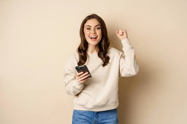 Happy brunette girl holding smartphone and cheering, winning, celebrating win on mobile phone app, standing over beige background — Stockfoto