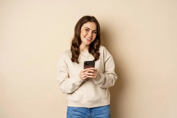 People and cellular technology. Beautiful stylish woman using mobile phone, smartphone app, smiling and looking at screen, standing over beige background — Stockfoto