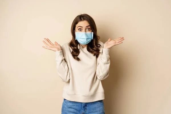 Covid-19, pandemic and quarantine concept. Surprised brunette woman in face medical mask, looking amazed at camera, watching big news, beige background — стоковое фото