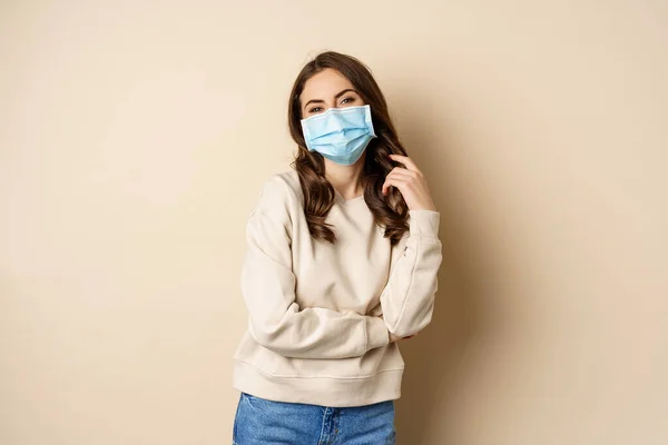 Covid-19, pandemic and quarantine concept. Beautiful modern woman in medical face mask, cross arms on chest, looking confident, beige background — ストック写真