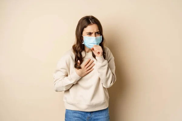 Covid-19和健康概念。头戴口罩的生病妇女咳嗽，喉咙酸痛，站在米黄色背景上 — 图库照片