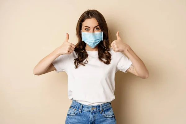 Covid-19 pandemic and health concept. Smiling happy woman showing thumbs up in medical face mask, standing over beige background — Fotografia de Stock