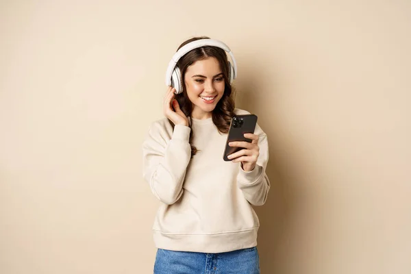 Beautiful young woman looking at video on smartphone, listening music in headphones, standing in casual clothes over beige background — ストック写真