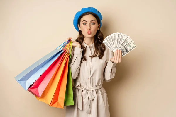 Stylish modern girl feeling satisfaction while shopping, posing with money dollars and shop bags, beige studio background — Foto de Stock