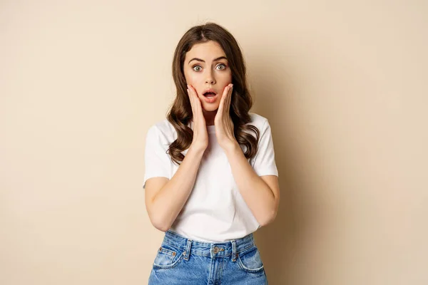 Portrait of stylish young woman gasping, looking surprised and amazed, impressed by smth, standing in t-shirt and jeans over beige background — Foto Stock