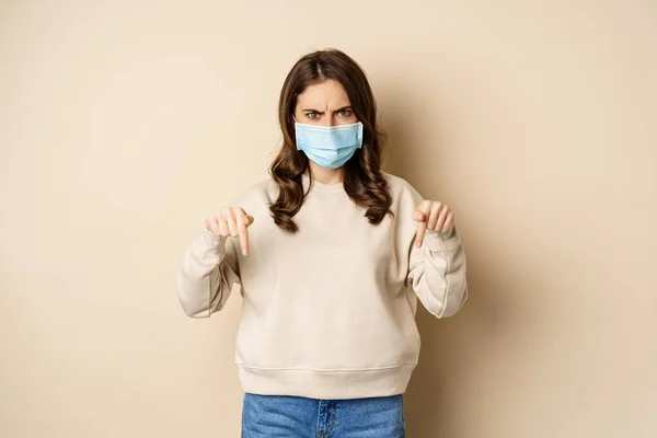 Angry woman in medical face mask, pointing fingers down and looking concerned, standing in sweater over beige background — стоковое фото