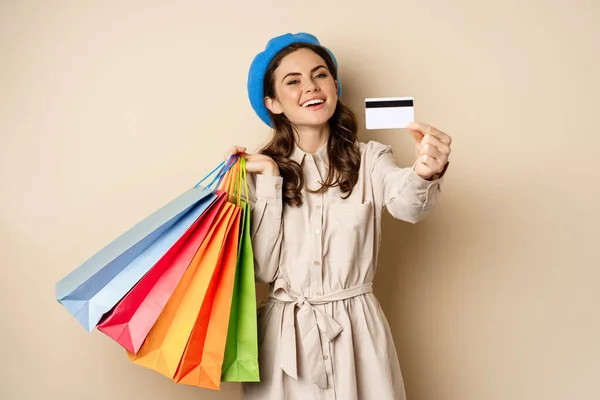 Portrat of trendy feminine girl posing with shopping bags from store and credit card, paying contactless, buying with discount on sale, beige background — Stock fotografie