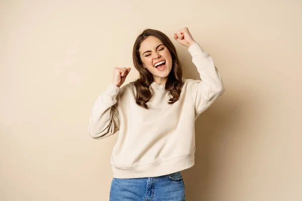 Enthusiastic brunette woman laughing and smiling, cheering, shaking hands and celebrating, triumphing, standing over beige background — Foto de Stock