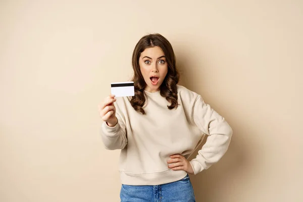 クレジットカードを示す驚きの女性,印象的な息をのむ,割引やショッピングの印象,ベージュの背景に立って — ストック写真