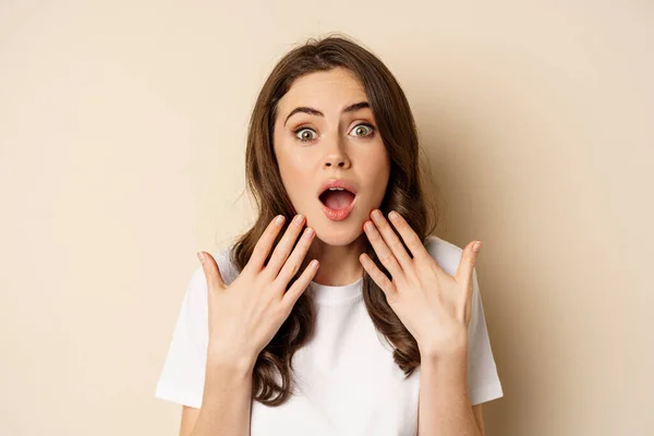 Close up portrait of surprised and excited brunette girl looking amazed, reacting impressed and excited, standing against beige background — стоковое фото