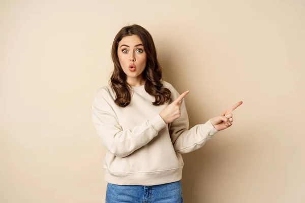 Sorpresa donna bruna indicando le dita a destra, mostrando pubblicità e dire wow impressionato, in piedi su sfondo beige — Foto Stock