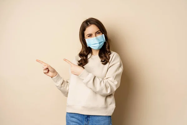 Health and covid-19 concept. Confused young woman in medical face mask, pointing left and staring with frustrated face, beige background — ストック写真