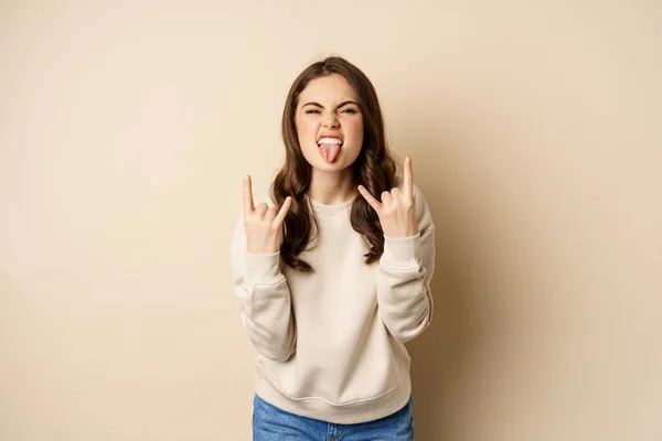 행복 한 젊은 여자가 음악을 즐기고, 즐거운 시간을 보내고, 록 음악을 연주하고, 헤비메탈 핑거 호른 몸짓을 하며, 베이지 색 배경 위에서 있다 — 스톡 사진