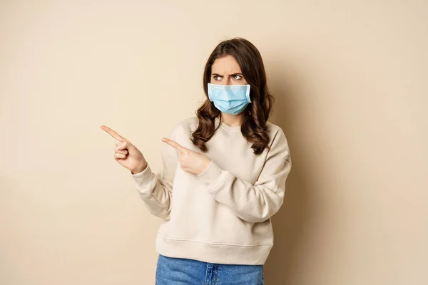 Health and covid-19 concept. Confused young woman in medical face mask, pointing left and staring with frustrated face, beige background — Photo