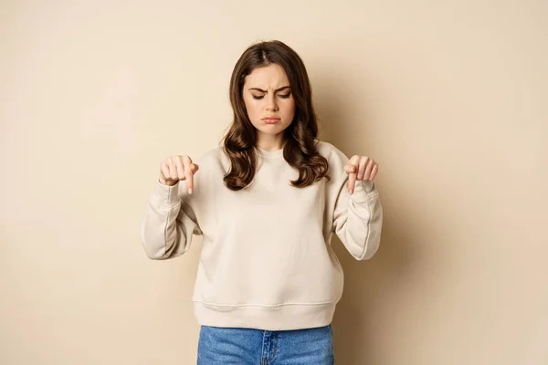 Sad and disappointed girl pointing, looking down with upset grimace moody face, standing over beige background — Foto de Stock
