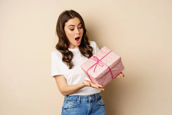 Concepto de celebración y festivos. Feliz joven mujer sosteniendo regalo envuelto en caja rosa, recibir regalo, mirando sorprendido y sorprendido, de pie sobre fondo beige — Foto de Stock