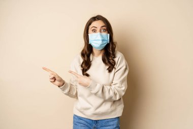 Sağlık ve covid-19 konsepti. Tıbbi maske takan heyecanlı bir kadın, parmaklarını sola gösteriyor ve şaşırmış görünüyor, promosyon teklifinden etkilenmiş, bej arka plan