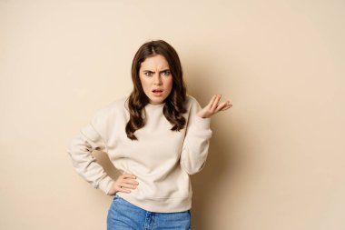 Frustrated and pissed-off woman, looking confused, arguing, standing over beige background
