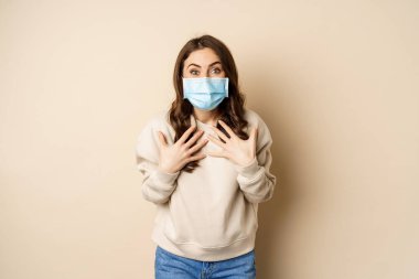 Covid-19, pandemic and quarantine concept. Surprised brunette woman in face medical mask, looking amazed at camera, watching big news, beige background
