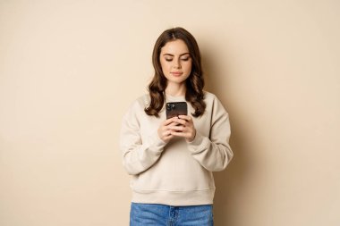İnsanlar ve hücresel teknoloji. Güzel stil sahibi bir kadın cep telefonu kullanıyor, akıllı telefon uygulaması kullanıyor, gülümsüyor ve ekrana bakıyor, bej arka planda duruyor.