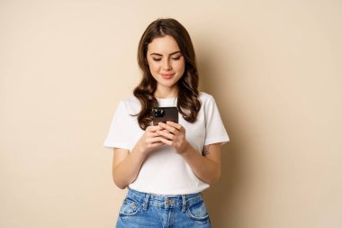 Genç modern kız uygulamayla sohbet ediyor, akıllı telefon uygulaması kullanıyor ve gülümsüyor, bej arka planda duruyor.