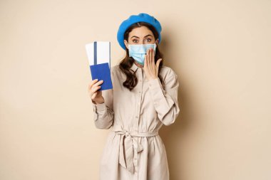 Covid pandemic and travelling concept. Portrait of cute girl in medical face mask going on trip, showing passport with tickets abroad and looking excited, beige background