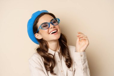 Close up portrait of stylish woman posing in sunglasses, laughing and smiling, wearing trendy outfit, standing over beige background