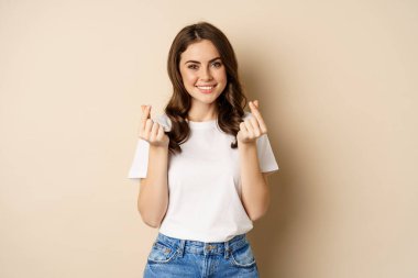Cute young woman showing finger hearts sign and smiling, express care and love, standing over beige background