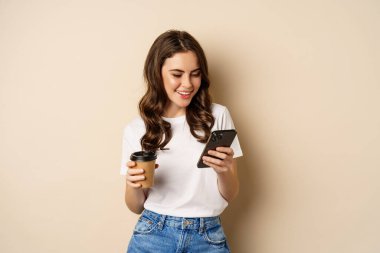 Smiling woman using mobile phone app and credit card, concept of online shopping, contactless payment and delivery, standing over beige background