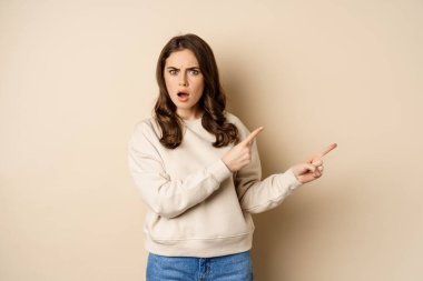 Shocked girl pointing fingers right, gasping startled, standing over beige background