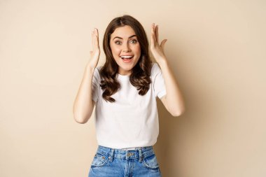 Enthusiastic adn surprised woman winning, dancing and celebrating, standing in tshirt over beige background