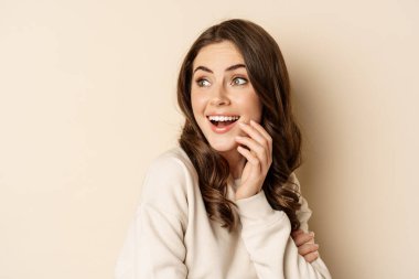 Close up portrait of brunette feminine woman turn back, look behind with surprised face expression, standing against beige background
