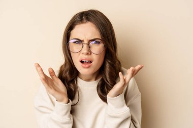 Close up portrait of woman in glasses looking confused and frustrated, cant understand smth strange, standing over beige background