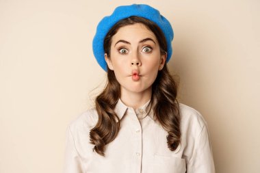 Young funny woman acting, fooling around, pucker lips and grimacing, posing against beige background in trendy blue hat