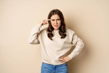 Frustrated woman roll finger near head and complaining, insulting someone, standing over beige background