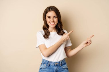 Smiling brunette woman in t-shirt, pointing fingers right, showing promo offer or advertisement, demonstrating banner, standing against beige background