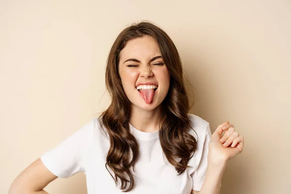 Close up of happy, carefree beautiful woman posing silly, showing tongue, having fun, standing in casual white t-shirt against beige background — ストック写真