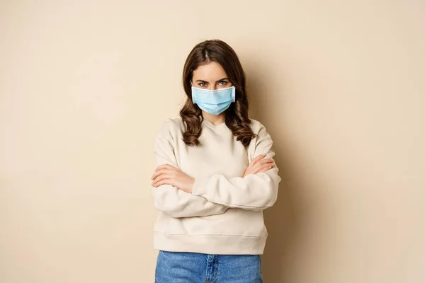 Covid-19, pandemic and quarantine concept. Beautiful modern woman in medical face mask, cross arms on chest, looking confident, beige background — Fotografia de Stock