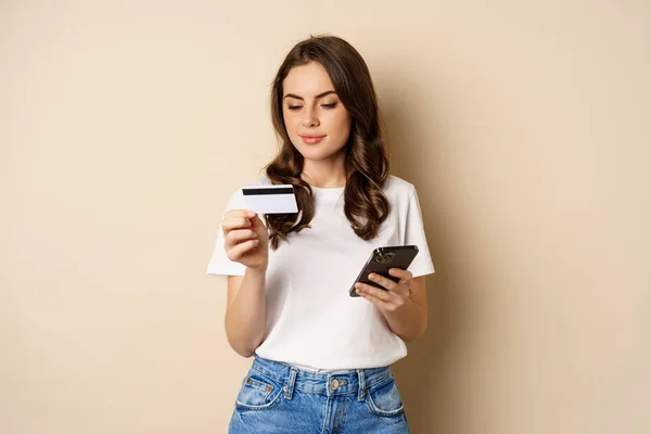 Image of young happy woman paying online, holding smartphone and credit card, enter info in application on mobile phone, standing against beige background — Foto de Stock