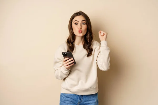 Happy girl holding smartphone and celebrating, winning on mobile phone app, standing over beige background — стоковое фото
