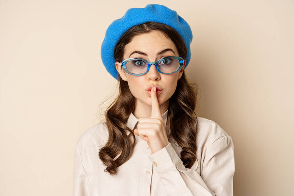 Secret. Young cute woman in sunglasses, shushing, showing taboo, hush sign, press finger to lips, standing over beige background