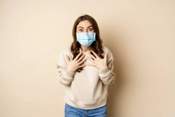 Covid-19, pandemic and quarantine concept. Surprised brunette woman in face medical mask, looking amazed at camera, watching big news, beige background — Photo