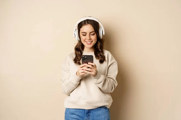 Sonriendo linda chica en los auriculares, mirando el teléfono móvil, escuchando música o podcast, de pie sobre fondo beige — Foto de Stock