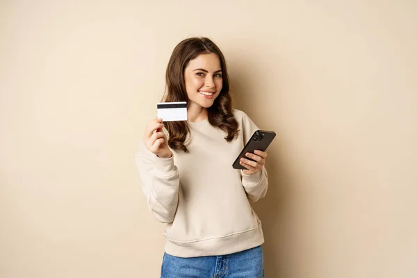 Online shopping. Smiling brunette woman showng credit card, using smartphone app, standing over beige background — Foto de Stock