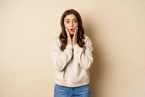 Image of surprised, amazed brunette girl gasping, looking fascinated at camera, standing over beige background — Stock fotografie